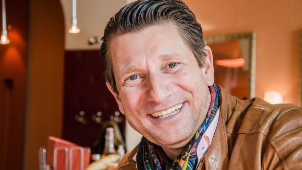 Ein lächelnder Mann mit brauner Lederjacke und buntem Schal.