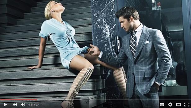 Eine Frau im Kleid sitzt auf einer Treppe, während ein Mann im Anzug sie berührt.