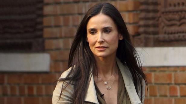 Demi Moore mit dunklen, langen Haaren und Halsketten vor einer Backsteinmauer.