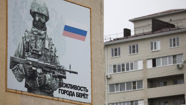 Ein Plakat zeigt einen Soldaten mit Gewehr und russischer Flagge vor einem Wohngebäude.