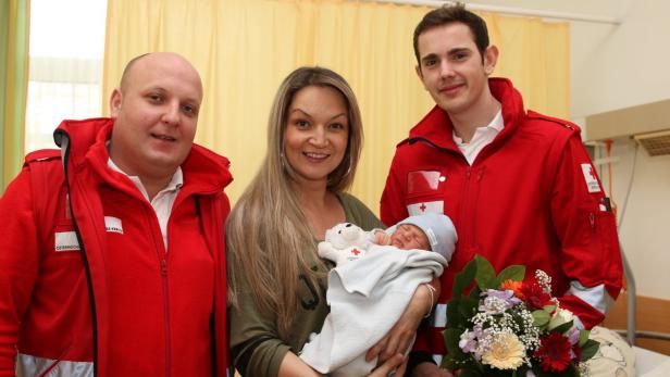 Eine Frau hält ein Neugeborenes im Arm, flankiert von zwei Sanitätern des Österreichischen Roten Kreuzes.