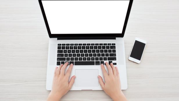 Eine Person tippt auf einem Laptop, daneben liegt ein Smartphone.