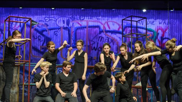 Eine Gruppe von Jugendlichen in schwarzen Kostümen steht auf einer Bühne vor einer Graffiti-Wand.
