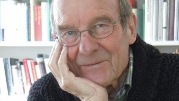 Ein älterer Mann mit Brille stützt sein Gesicht in die Hand vor einem Bücherregal.