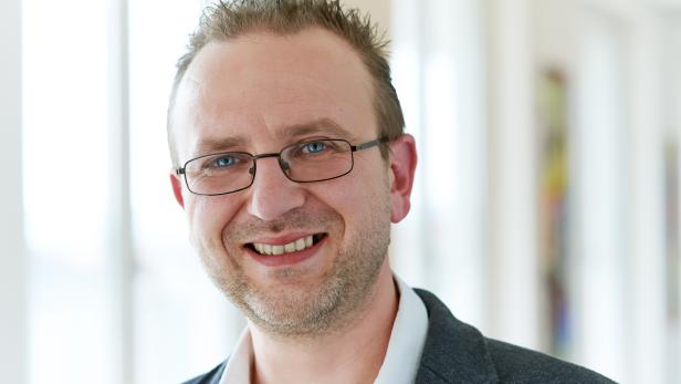 Ein lächelnder Mann mit Brille und grauem Sakko vor hellem Hintergrund.