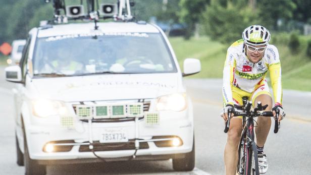 Ein Radfahrer fährt auf einer Straße, begleitet von einem Begleitfahrzeug.
