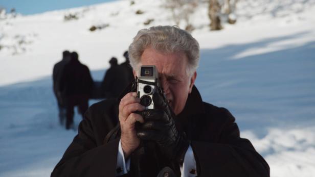 Ein Mann mit grauen Haaren fotografiert mit einer alten Kamera in einer verschneiten Landschaft.