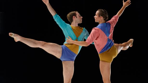 Zwei Tänzer in farbenfrohen Kostümen führen eine Ballettpose vor schwarzem Hintergrund auf.