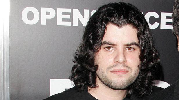 Ein Mann mit lockigem, dunklem Haar vor einer Wand mit der Aufschrift „OPEN“.