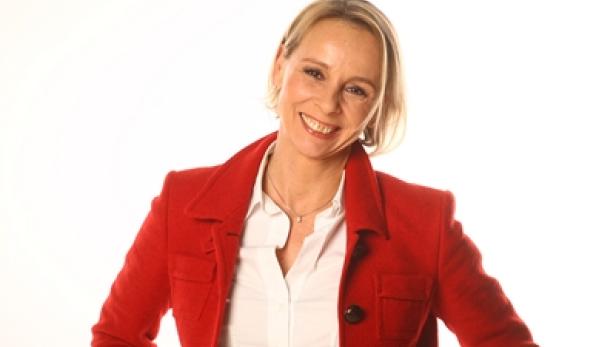 Eine lächelnde Frau mit blonden Haaren trägt ein rotes Jackett und eine weiße Bluse.