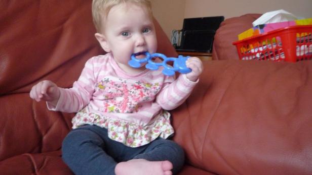 Ein blondes Baby sitzt auf einem Sofa und kaut auf einem blauen Spielzeug herum.