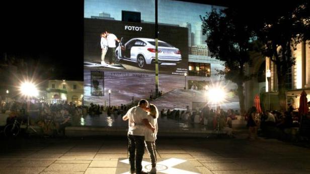 Ein Paar umarmt sich vor einer Projektion mit einem Auto und dem Schriftzug „Foto?“.
