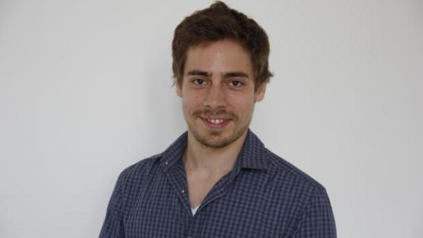 Ein junger Mann mit braunen Haaren und einem karierten Hemd lächelt.