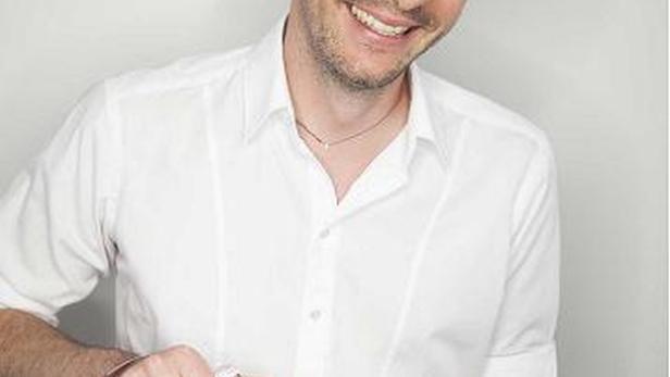 Ein lächelnder Mann im weißen Hemd hält eine Flasche Almdudler.