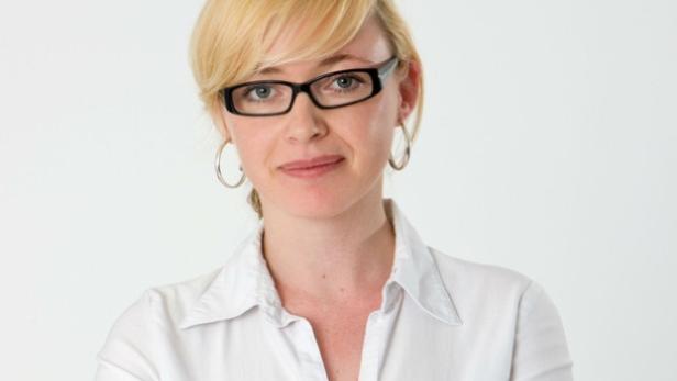 Eine blonde Frau mit Brille und weißem Hemd lächelt.