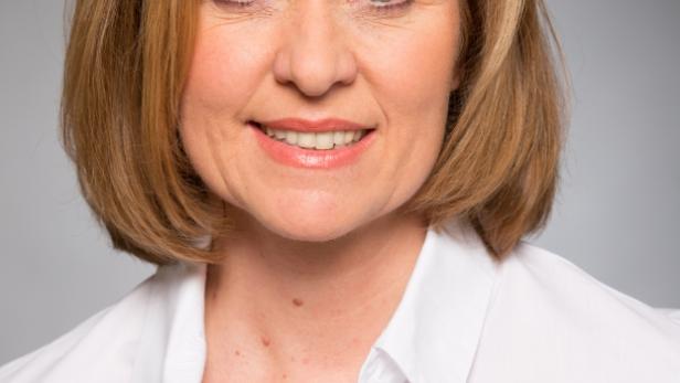 Porträt einer lächelnden Frau mit blonden Haaren und einem weißen Hemd vor grauem Hintergrund.