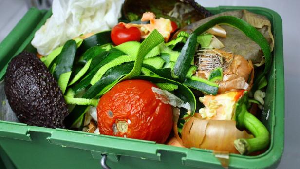 Ein grüner Kompostbehälter gefüllt mit organischen Abfällen wie Avocado, Tomaten und Zucchini.