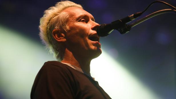 Ein Mann mit blonden Haaren singt in ein Mikrofon auf einer Bühne.