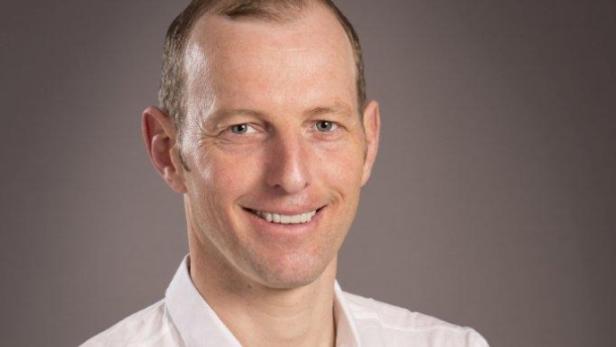 Ein lächelnder Mann mit kurzem, blondem Haar vor einem grauen Hintergrund.