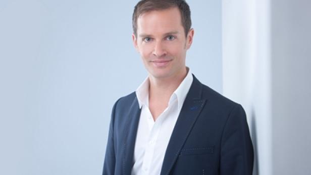 Ein Mann in einem blauen Sakko und einem weißen Hemd lehnt an einer Wand.