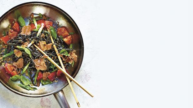 Eine Wok-Pfanne mit schwarzen Nudeln, Tomaten, Trüffeln und Basilikum, serviert mit Essstäbchen.