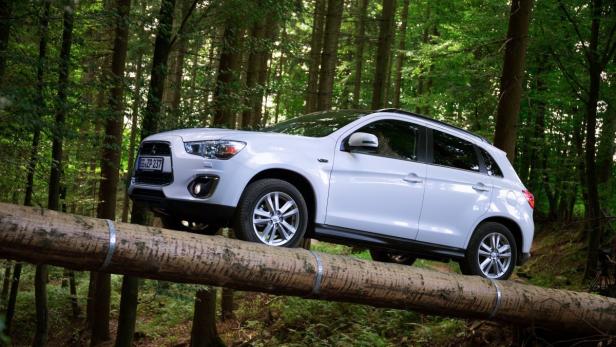 Ein weißer Mitsubishi fährt über einen Baumstamm im Wald.