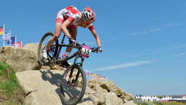 Eine Mountainbikerin fährt über eine felsige Strecke mit verschiedenen Nationalflaggen im Hintergrund.