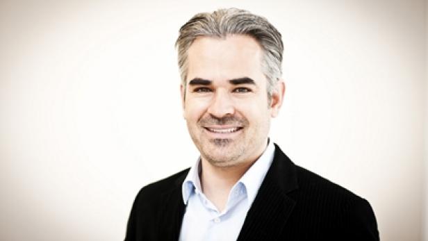 Ein Mann mit grau meliertem Haar lächelt in einem schwarzen Sakko.