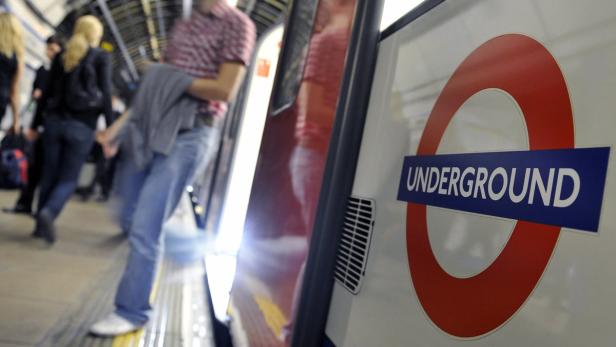 Das Logo der Londoner U-Bahn an einem Bahnsteig mit wartenden Menschen.