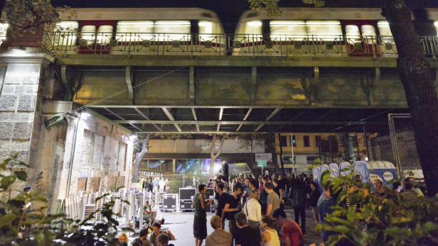 Eine Menschenmenge unter einer Eisenbahnbrücke bei Nacht, während ein Zug darüberfährt.