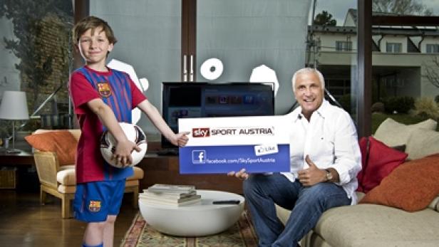 Ein Junge im Fußballtrikot und ein Mann halten ein Schild von Sky Sport Austria in einem Wohnzimmer.