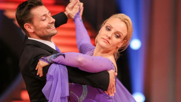 Ein Tanzpaar in lila Kleidung tanzt vor einem roten und blauen Hintergrund.