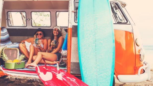 Zwei junge Frauen sitzen in einem VW-Bus mit Surfbrettern am Strand und trinken Smoothies.