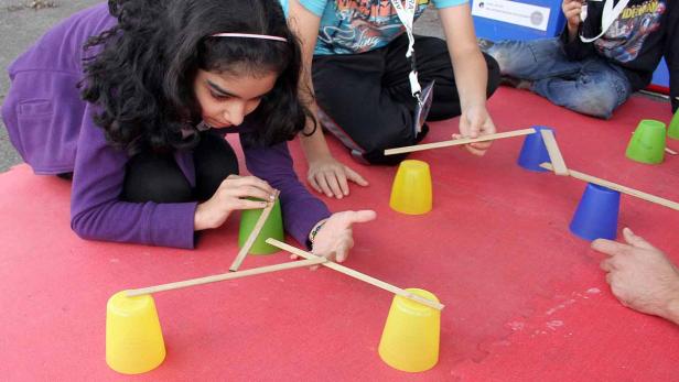 Kinder balancieren Holzstäbe auf Plastikbechern.