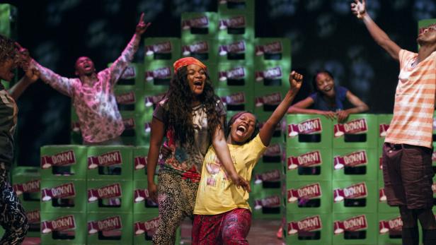 Eine Gruppe von Menschen tanzt und jubelt auf einer Bühne vor Getränkekisten.