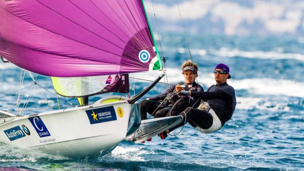 Zwei Segler steuern ein Segelboot mit einem lila Segel auf dem offenen Meer.