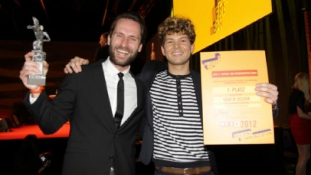 Zwei Männer posieren mit einer Trophäe und einer Urkunde für „1. Platz Grafik Design“.