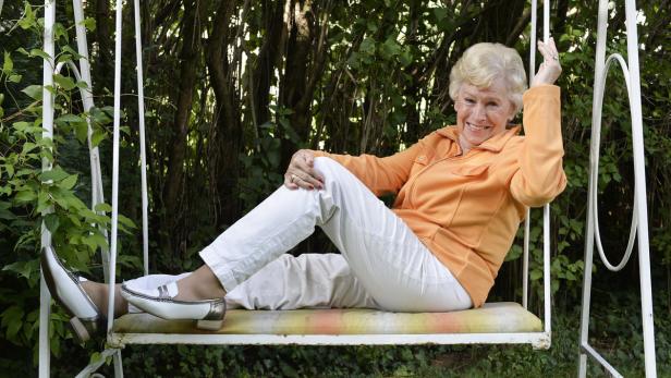 Eine ältere Frau sitzt lächelnd auf einer Gartenschaukel.