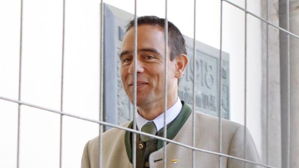Ein lächelnder Mann in Trachtenjacke steht hinter einem Metallzaun.