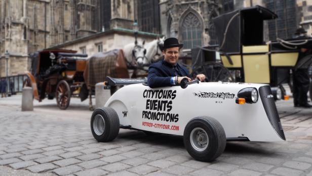 Ein Mann mit Melone fährt ein Hot-Rod-Auto in Wien, im Hintergrund Pferdekutschen.