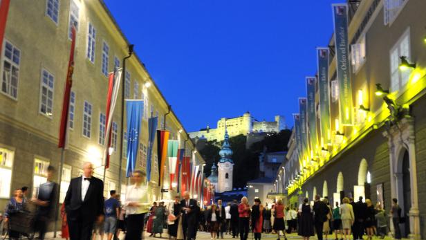 Menschen flanieren abends in Salzburg, geschmückt mit Flaggen und Bannern, im Hintergrund die Festung Hohensalzburg.