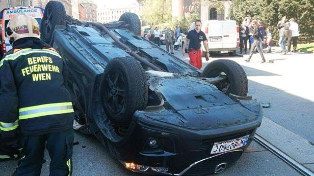 Ein auf dem Dach liegendes Auto in Wien, umgeben von Feuerwehrleuten und Zuschauern.