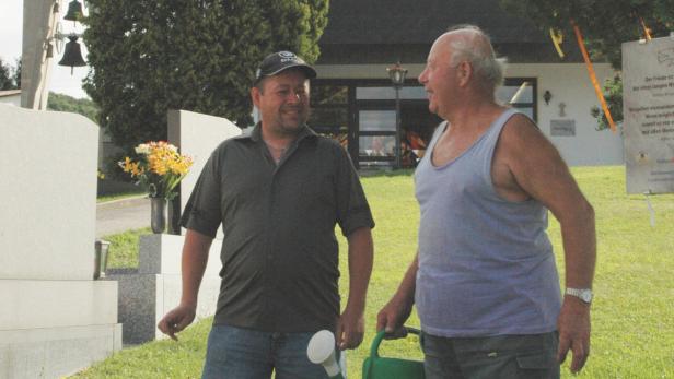 Zwei Männer unterhalten sich auf einem Friedhof, einer hält eine Gießkanne.