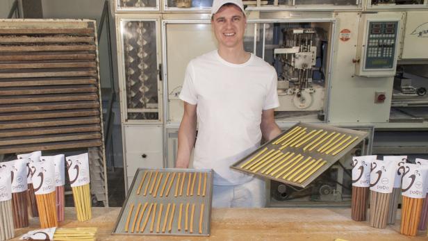 Ein Bäcker präsentiert in einer Backstube Bleche mit Teigstangen und verpackte Produkte.