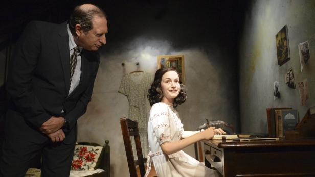 Ein Mann im Anzug betrachtet Anne Frank, die an einem Schreibtisch sitzt und schreibt.