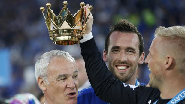 Ein Fußballspieler hält eine goldene Krone über dem Kopf, während zwei Männer lächeln.