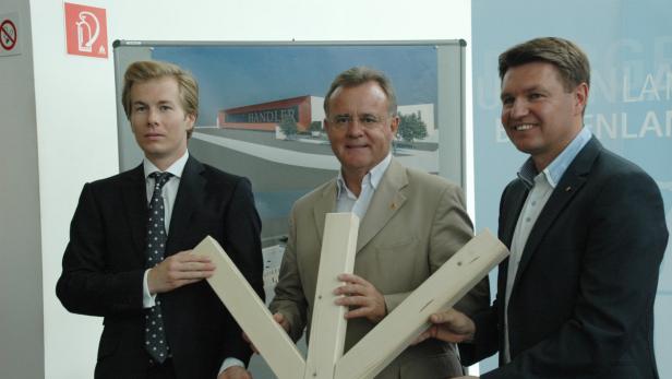 Drei Männer präsentieren ein Holzmodell vor einem Bild des „Handler“-Gebäudes.