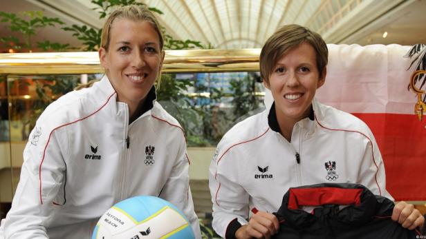 Zwei österreichische Beachvolleyballspielerinnen posieren mit einem Ball und einer Jacke vor einer Österreich-Flagge.