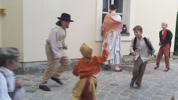 Eine Gruppe von Kindern in historischen Kostümen spielt auf einem gepflasterten Platz.