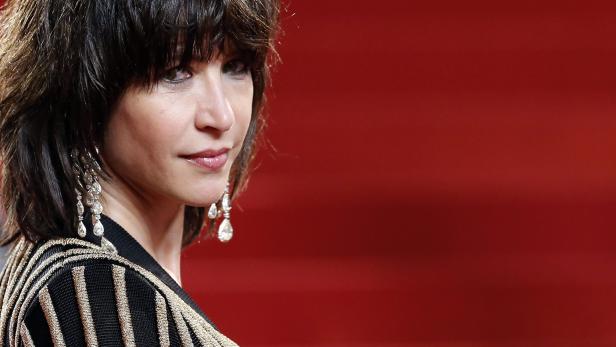 Sophie Marceau mit dunklen Haaren und auffälligen Ohrringen vor rotem Hintergrund.